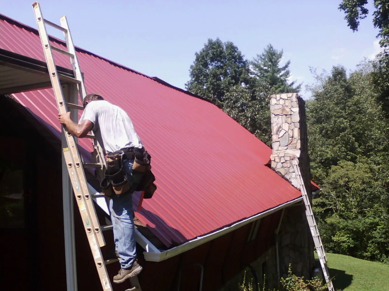 Roofer on a ladder beside a red metal roof and stone chimney — Soffit & Fascia Repair by Roofing Brothers in Earth City